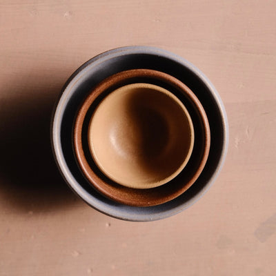 Overhead view of three nested Settle Ceramics Wander Side Bowls in grey, reddish brown and golden tan, showing their contrasting glazes and concentric rims on a brown tabletop.