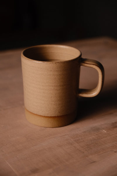 Tan Settle Ceramics Wander Cappuccino Mug on a brown tabletop, viewed at an angle to show its cylindrical ceramic body, rounded rectangular handle, fine speckles and contrasting ochre band around the base.