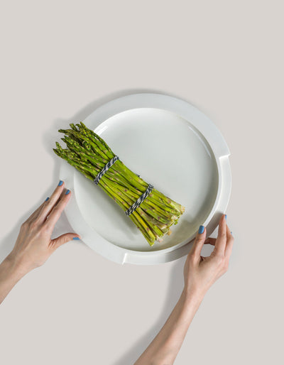 Overhead view of Tortuga Forma Kaolin Platter White holding a bundle of green asparagus tied with dark cord, with two hands with blue-painted nails holding its edges, showing a shallow round centre and broad rim divided by stepped notches.