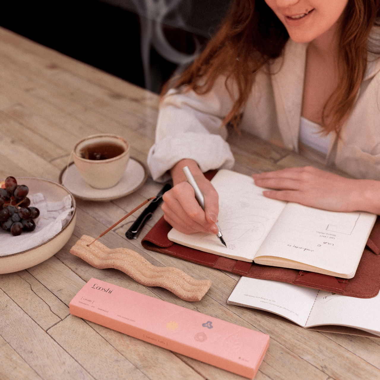 Woman writing in an open notebook at a wooden table with coffee and a peach box of Looshi Create incense sticks nearby