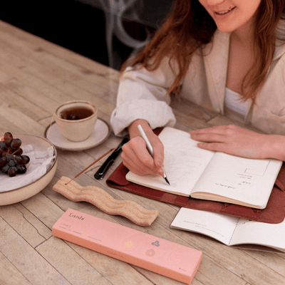 Woman writing in an open notebook at a wooden table with coffee and a peach box of Looshi Create incense sticks nearby
