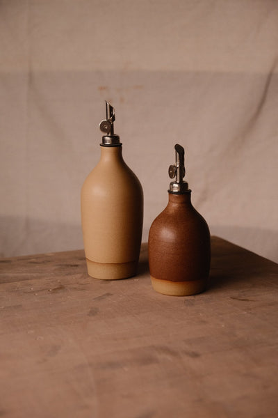 Rust-brown Settle Ceramics Wander Oil Bottle beside a taller golden-tan bottle on a wooden tabletop, both fitted with metal pouring spouts and casting long shadows against a beige backdrop.