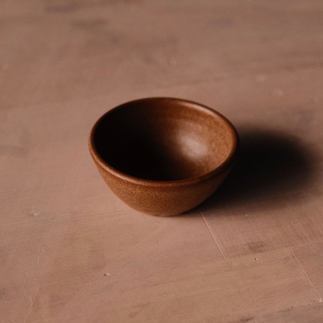 Warm reddish-brown Settle Ceramics Wander Pinch Bowl viewed at an angle on a brown tabletop, showing its rounded ceramic body, gently flared rim and subtly speckled glaze, with a dark shadow extending right.