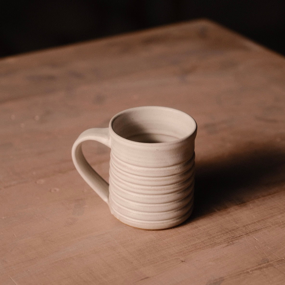 Cream Settle Ceramics The Little Mug viewed slightly from above on a wooden tabletop, showing its cylindrical ceramic body, pronounced horizontal ridges, curved handle on the left and open interior, with a long shadow extending right.