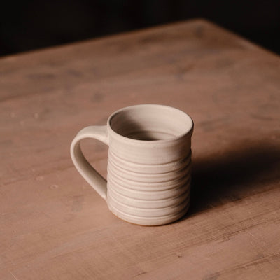 Cream Settle Ceramics The Little Mug viewed slightly from above on a wooden tabletop, showing its cylindrical ceramic body, pronounced horizontal ridges, curved handle on the left and open interior, with a long shadow extending right.