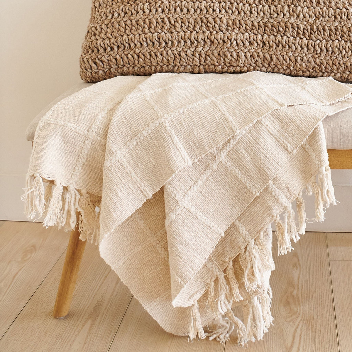 Variously Sea Shell White handloom throw draped over a pale wood bench, showing its raised check pattern, soft texture and thick knotted fringe