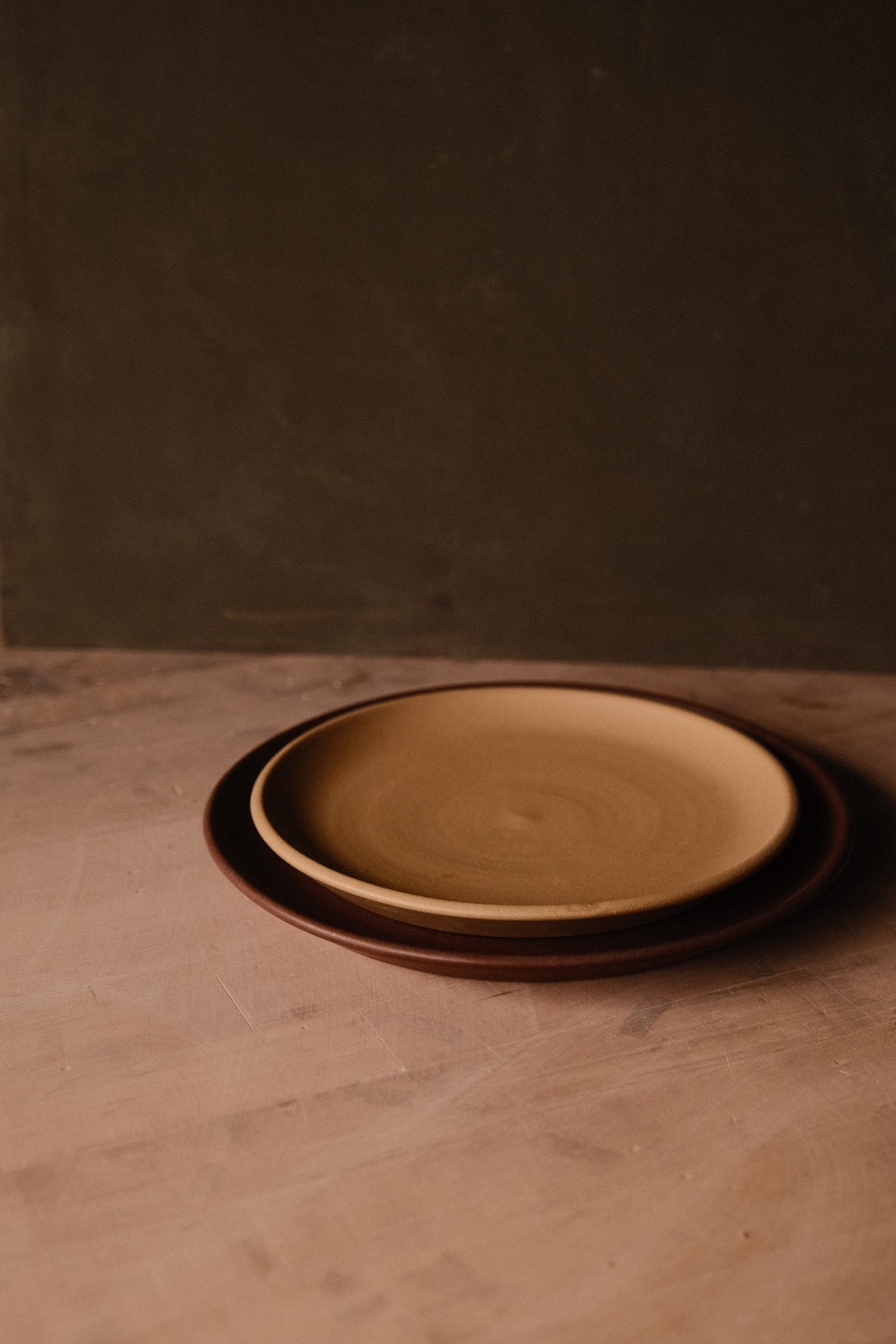 Golden-tan Settle Ceramics Horizon Lunch Plate resting on a larger dark brown plate on a wooden tabletop, viewed at a low angle to show its shallow profile and raised rim against a dark backdrop.