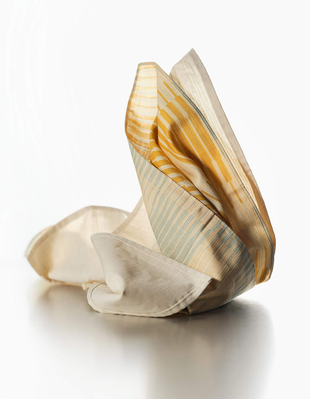 Tortuga Forma Dinner Napkins Double Sided Drift - Quiet Season gathered into upright folds on a light surface, revealing contrasting cream, mustard-yellow and pale grey-green patterned sides and fine woven texture.
