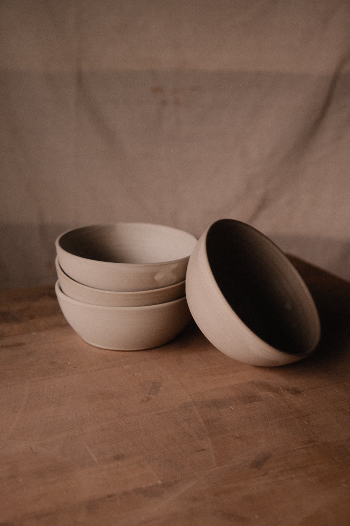 Cream Settle Ceramics Wander Soup Bowls arranged in a stack of three with another tilted alongside, revealing their rounded profiles and shadowed interiors on a wooden tabletop against beige fabric.