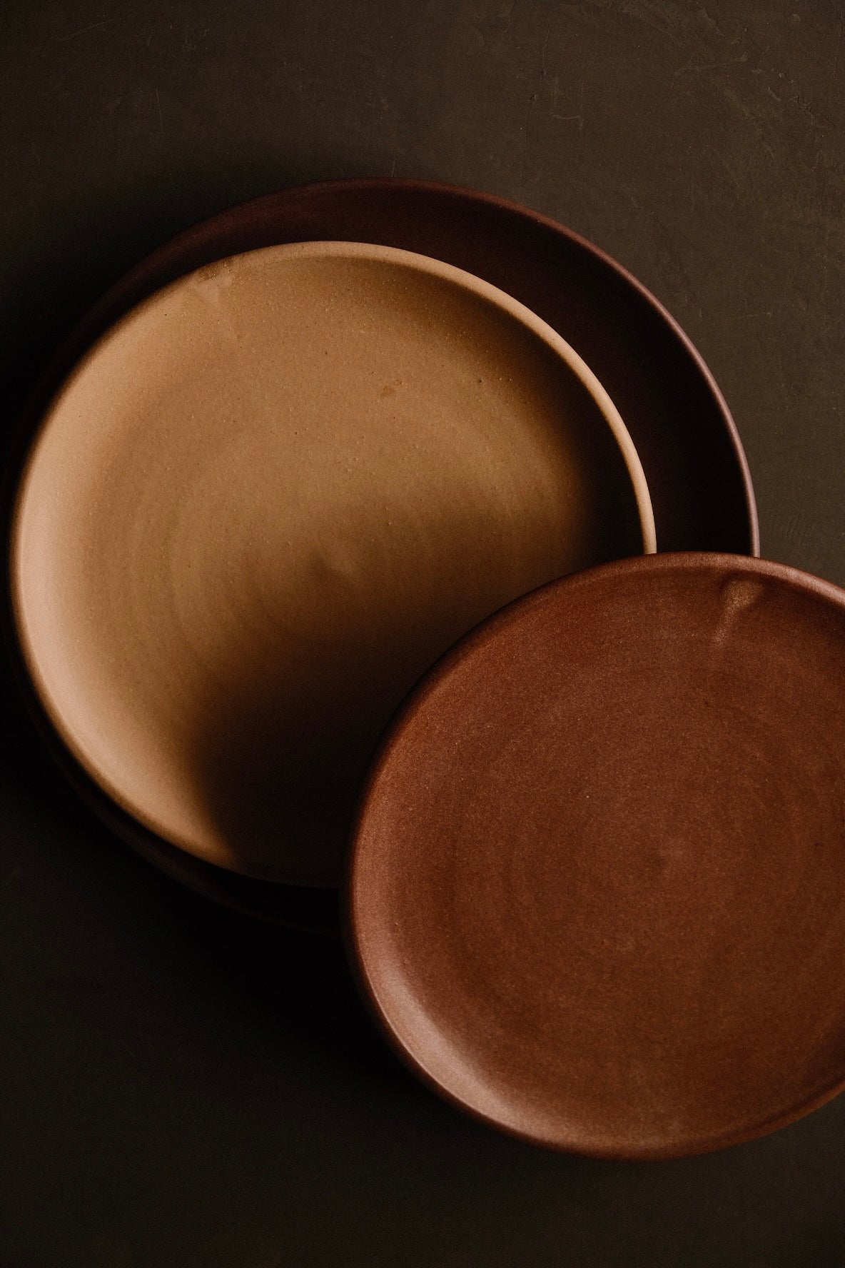 Overhead view of overlapping Settle Ceramics Horizon Lunch Plates in golden tan, reddish brown and dark brown on a dark surface, showing round ceramic forms, gently raised rims and subtle concentric surface lines.