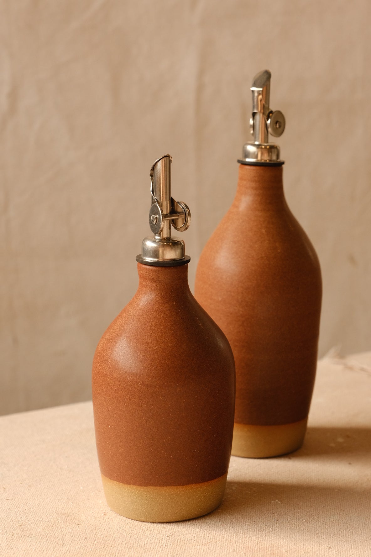 Rust-orange Settle Ceramics Wander Tall Oil Bottle beside a shorter matching bottle, both with finely speckled glaze, rounded shoulders, metal pourers and pale tan base bands.