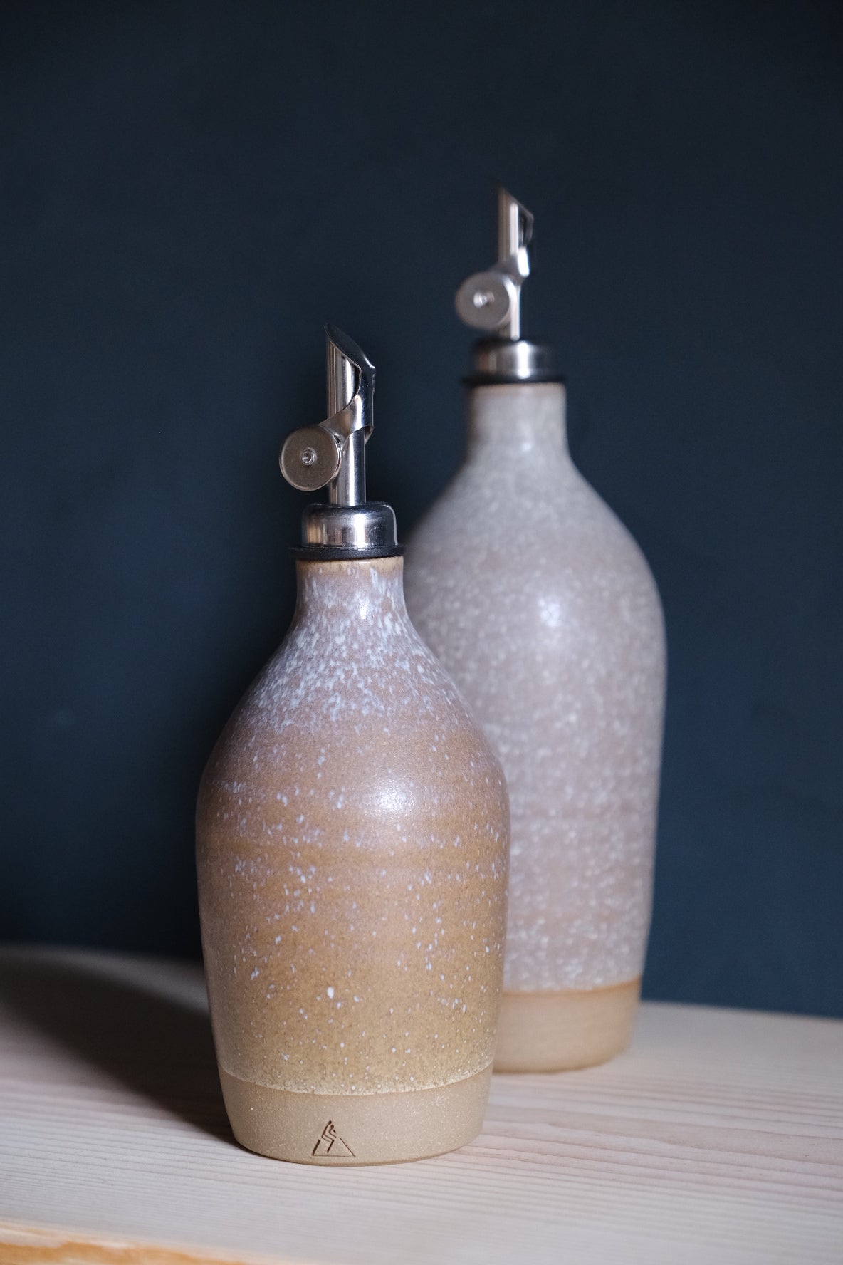 Pale lavender-grey Settle Ceramics Wander Tall Oil Bottle behind a shorter tan bottle with white speckling, both fitted with silver-toned pouring spouts on a light wooden surface.