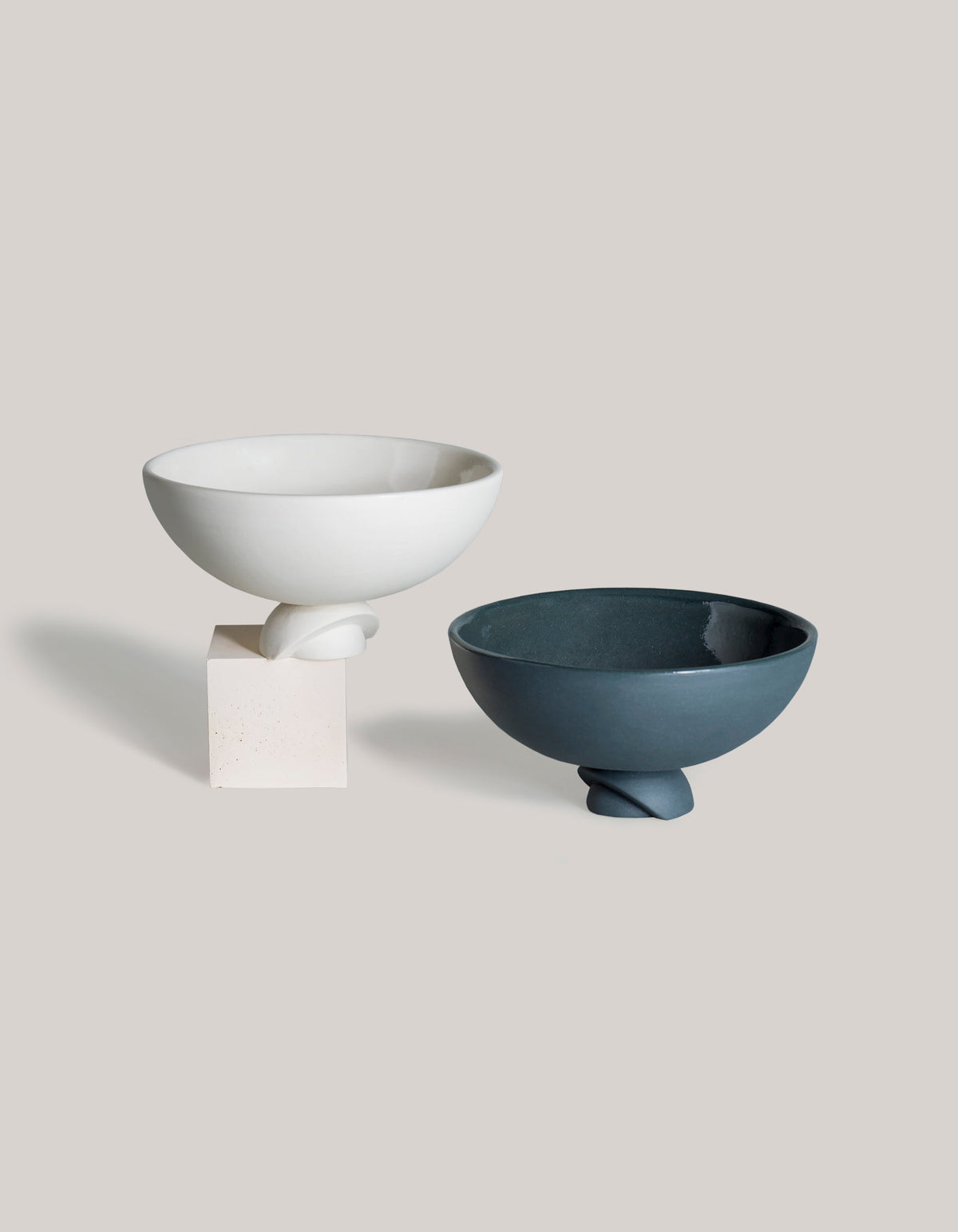 White and graphite Tortuga Forma Kaolin Rice Bowls displayed together against light grey, with the white bowl raised on a cream block and the graphite bowl resting beside it, showing their rounded bodies and sculptural feet.