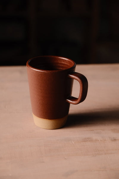 Full view of a reddish-brown Settle Ceramics Wander Latte Mug with its handle facing right and sandy-beige base visible, casting a long shadow across a wooden tabletop against a dark background.