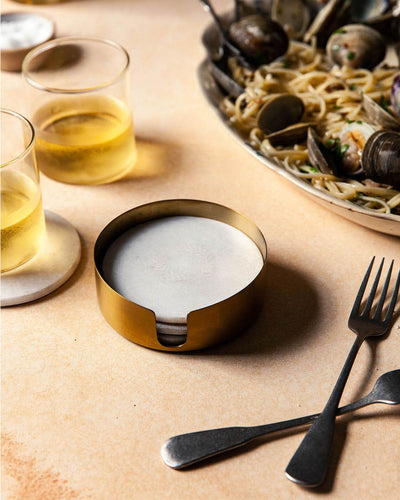 Brass holder of white marble coasters arranged on a dining table beside golden drinks, dark flatware, and a platter of food.