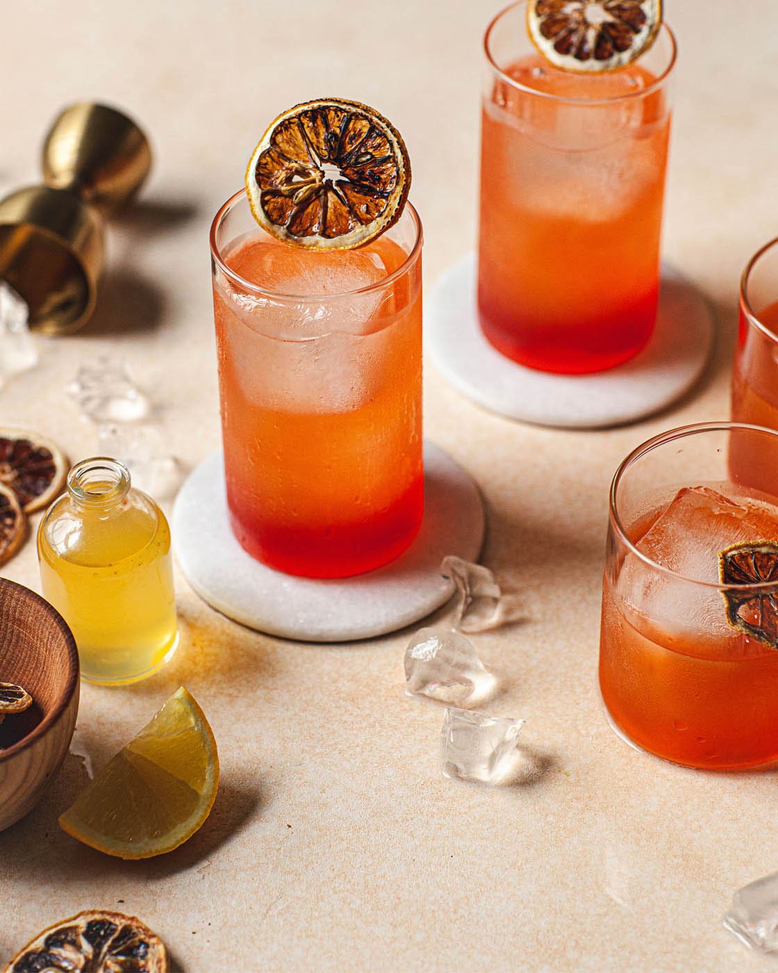 Red-orange cocktails resting on round white marble coasters, styled with dried citrus slices, scattered ice, and cocktail-making ingredients.