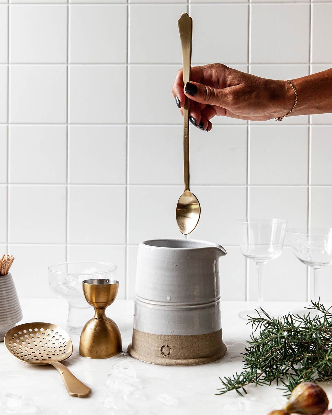 Hand holding the long gold bar spoon above a white ceramic mixing vessel, with the matching jigger and strainer nearby.