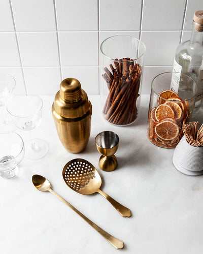 Essex gold barware set arranged on a marble counter beside cocktail glasses, bitters, dried citrus slices, and bar accessories.