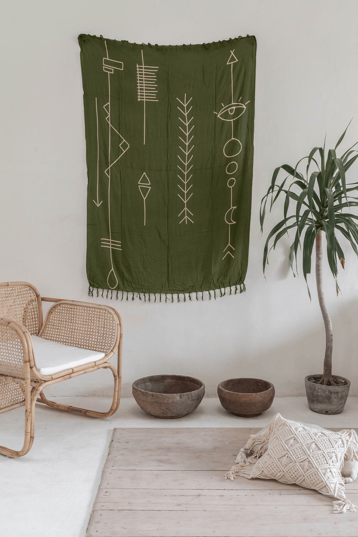 Village Thrive Abstract Tapestry, olive green rayon, hanging above a rattan chair with a potted plant and floor cushion.