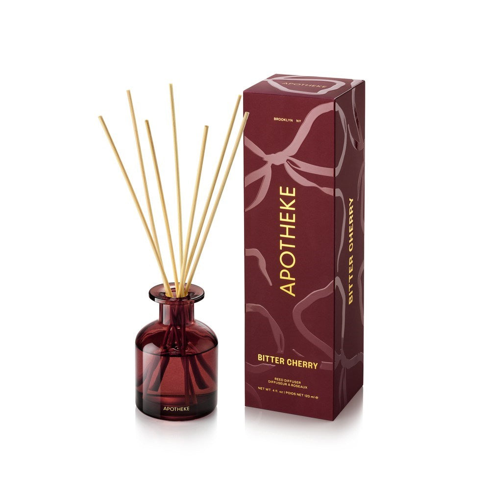 APOTHEKE Bitter Cherry Mini Reed Diffuser in a deep burgundy glass bottle with fanned natural-colored reeds, beside a tall burgundy patterned box with gold lettering on a white background.