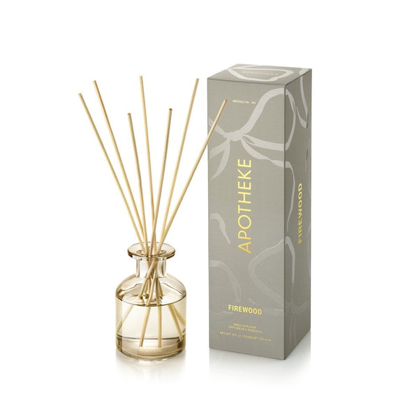 APOTHEKE Firewood Mini Reed Diffuser in a small clear glass bottle holding pale golden liquid and fanned natural-colored reeds, beside a tall gray-green patterned box with gold lettering.