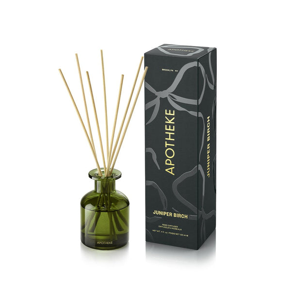 APOTHEKE Juniper Birch Mini Reed Diffuser in a green glass bottle with fanned natural-colored reeds, beside a tall dark green patterned box with gold lettering on a white background.