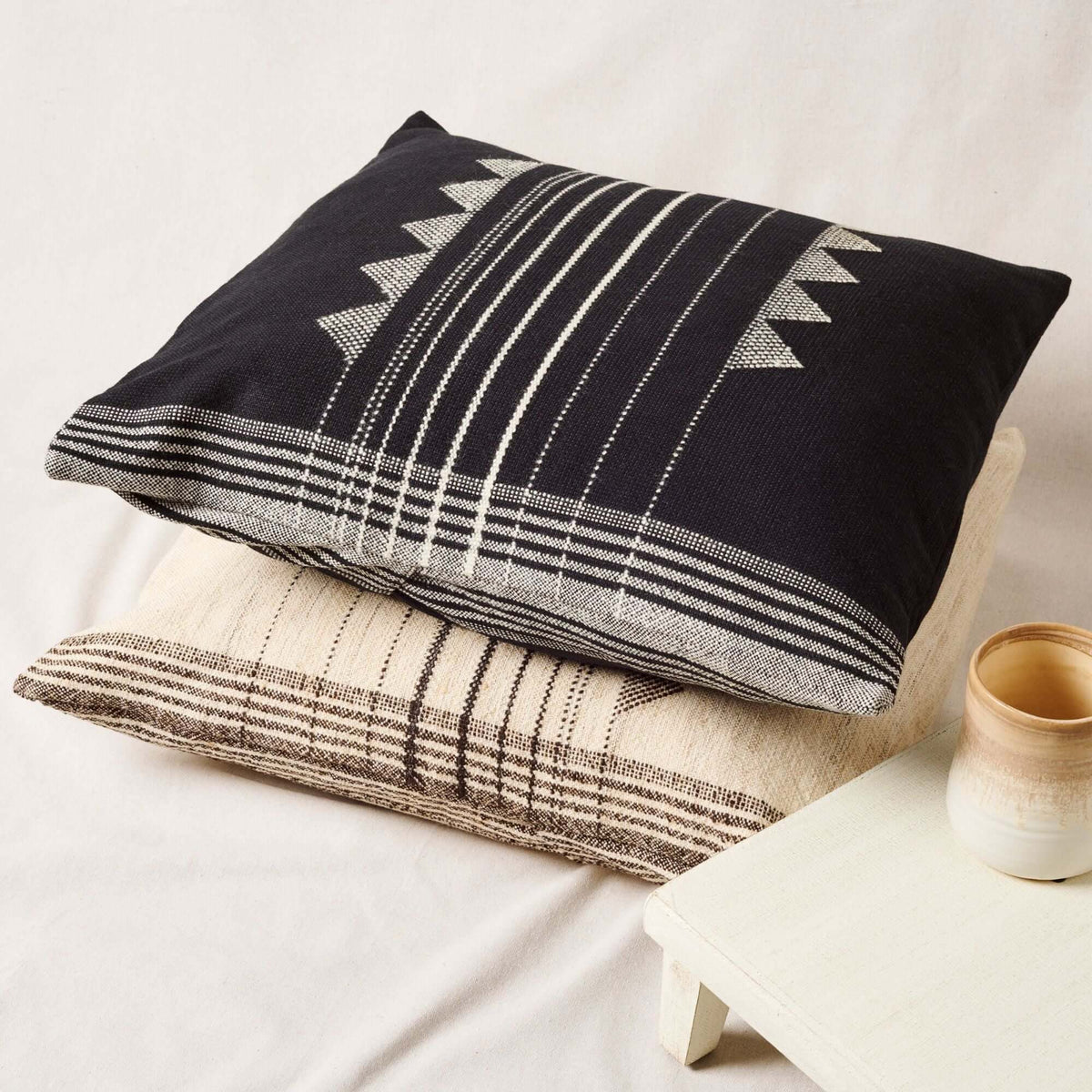 Kora Black handloom pillow stacked over a neutral plaid cushion beside a cup and open book
