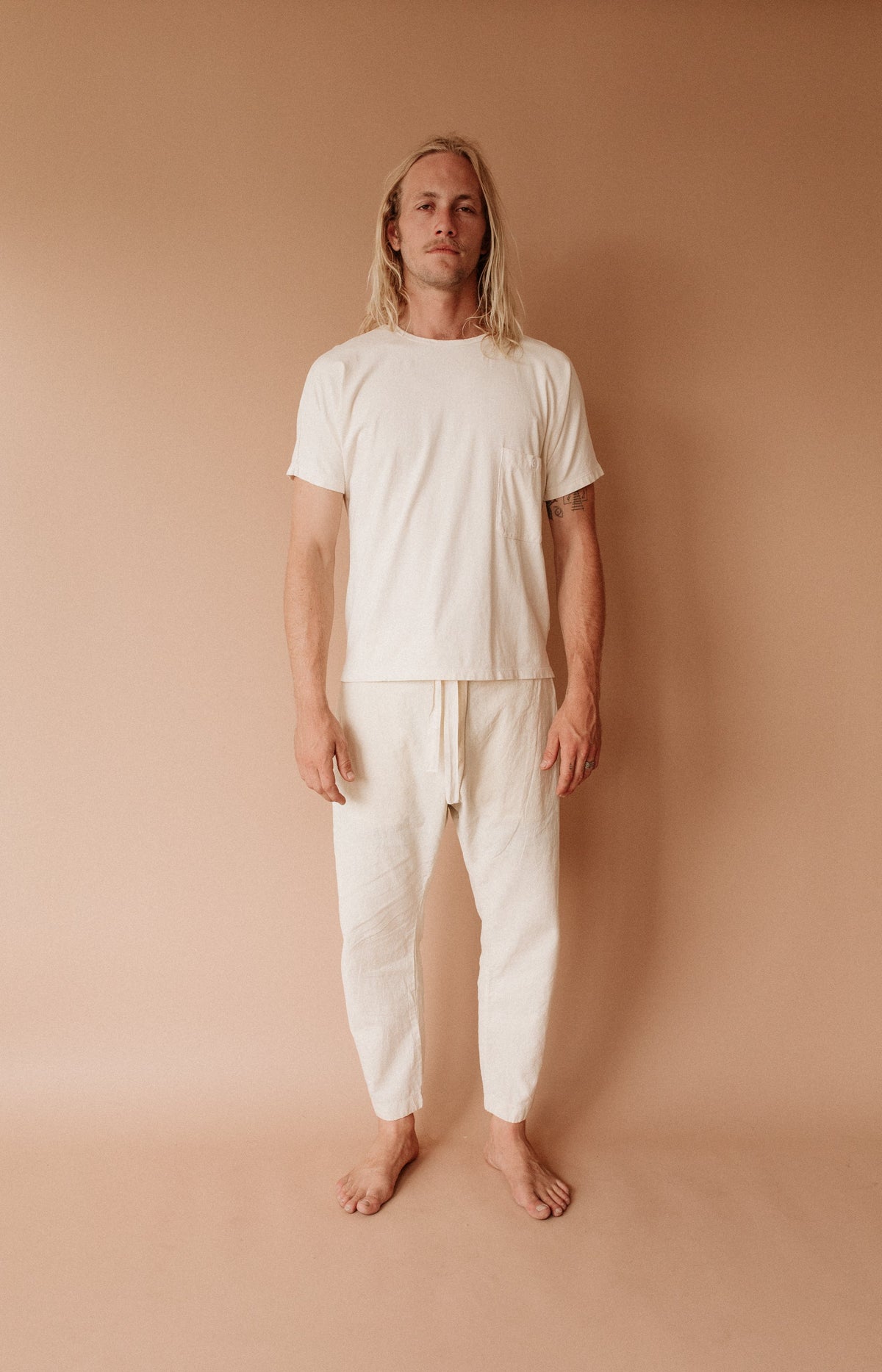 Barefoot model wearing a cream Pocket Dolman Tee with matching cropped pants against a warm tan backdrop.