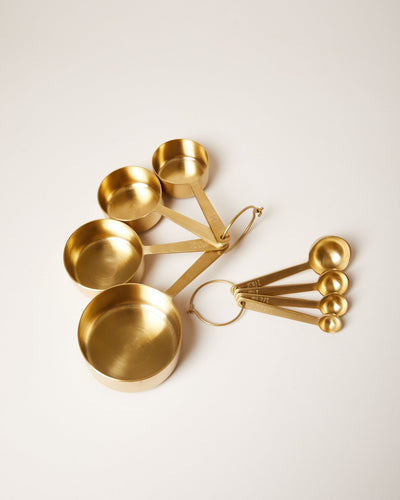 Coordinating brushed brass measuring cup and measuring spoon sets displayed side by side on a pale surface.