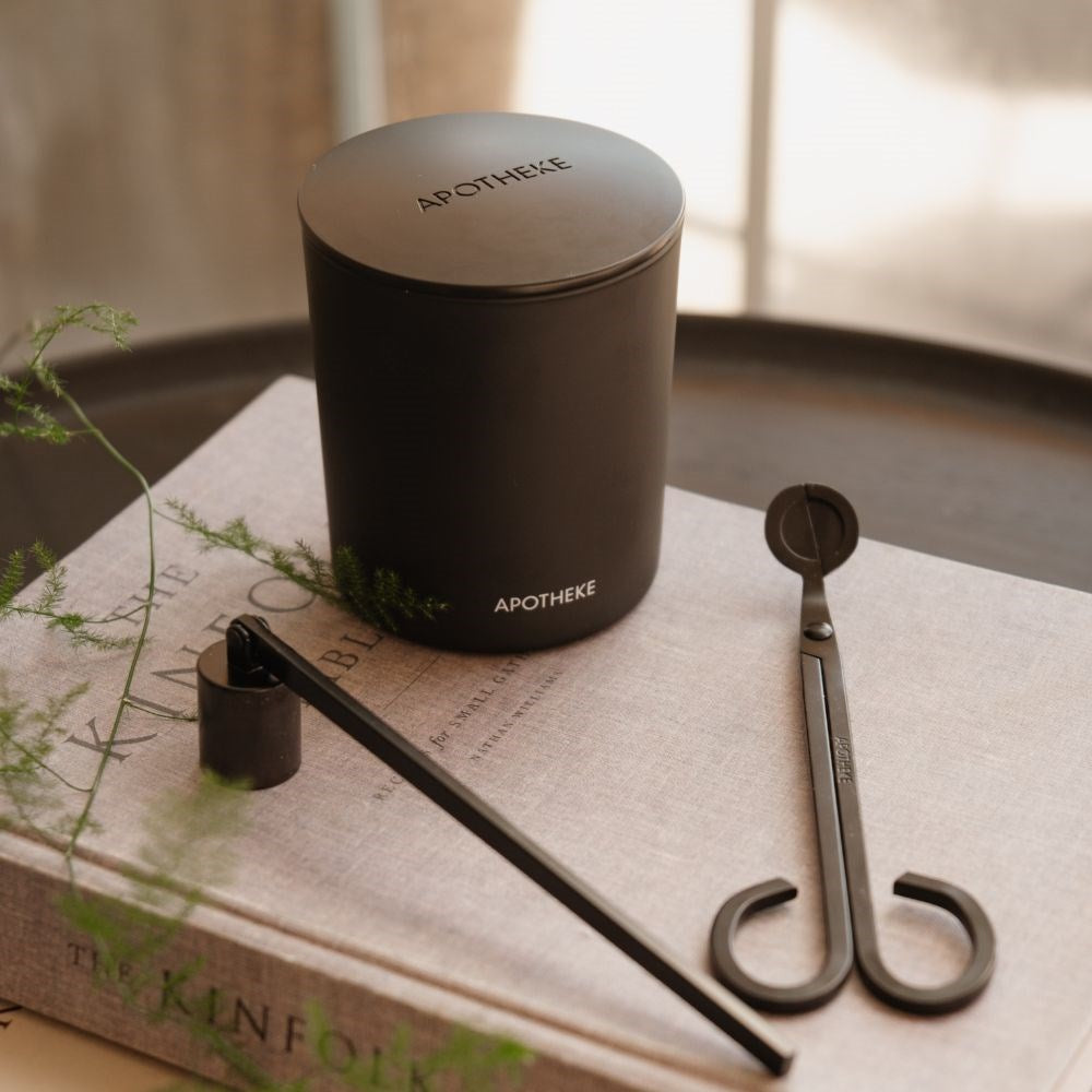Black wick trimmer and long-handled candle snuffer resting on a pale book beside a lidded black APOTHEKE candle, with greenery and soft window light.