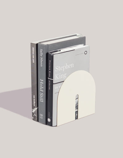 Cream bookend from Tortuga Forma Dumbo Bookend Pairs - The Architect Collection supporting three upright books on a light grey surface, showing its arched front and narrow slot with book edges visible through it.