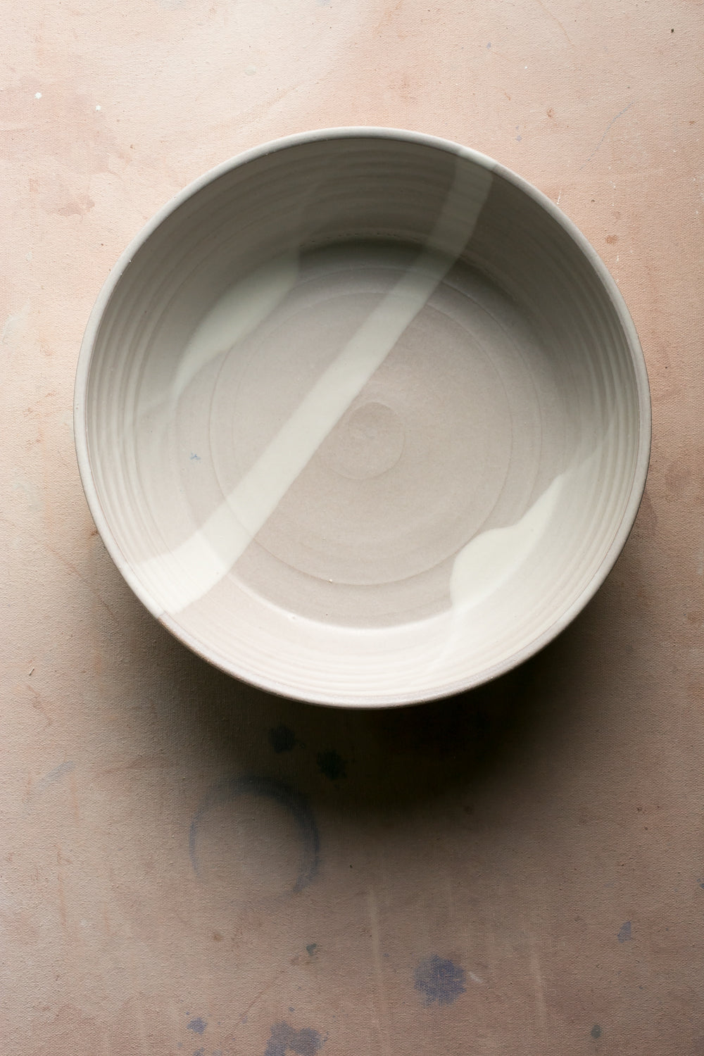 Overhead view of a pale grey-green Settle Ceramics Wander Serving Bowl on a tan tabletop, showing its broad interior, concentric surface rings and a diagonal stripe of sunlight.