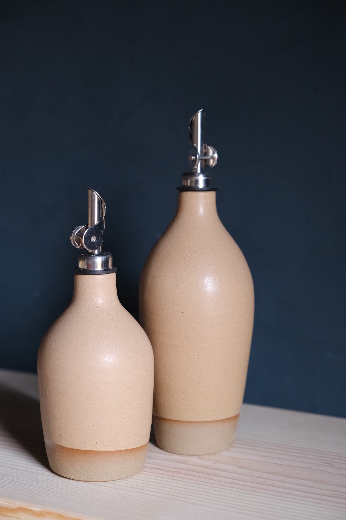 Cream Settle Ceramics Wander Oil Bottle in front of a taller pale blush-beige bottle on pale wood against a dark blue background, showing glossy rounded bodies, metal pouring spouts and light tan bases.