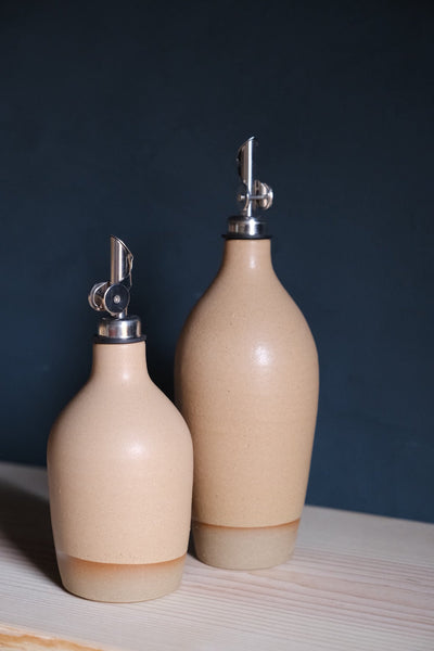 Cream Settle Ceramics Wander Oil Bottle in front of a taller pale blush-beige bottle on pale wood against a dark blue background, showing glossy rounded bodies, metal pouring spouts and light tan bases.