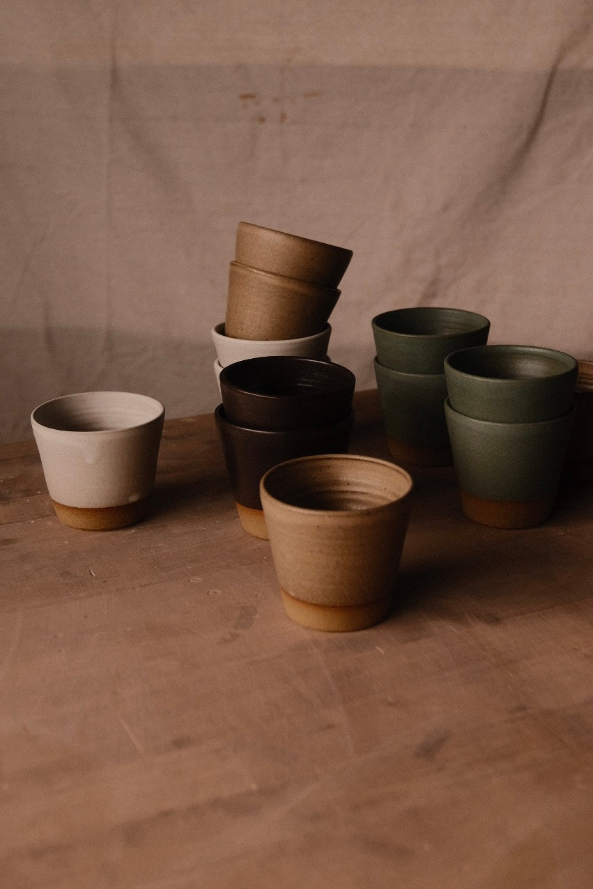 Settle Ceramics Wander Tumblers in cream, golden tan, dark brown and green arranged singly and in tilted stacks on a wooden tabletop against beige fabric, showing round openings, subtly ridged sides and ochre bases.