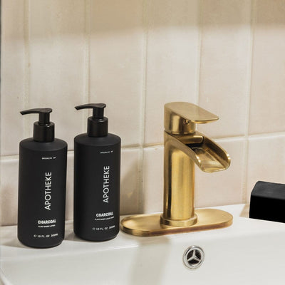 APOTHEKE Charcoal Lotion and Liquid Soap bottles styled on a bathroom sink beside a gold faucet
