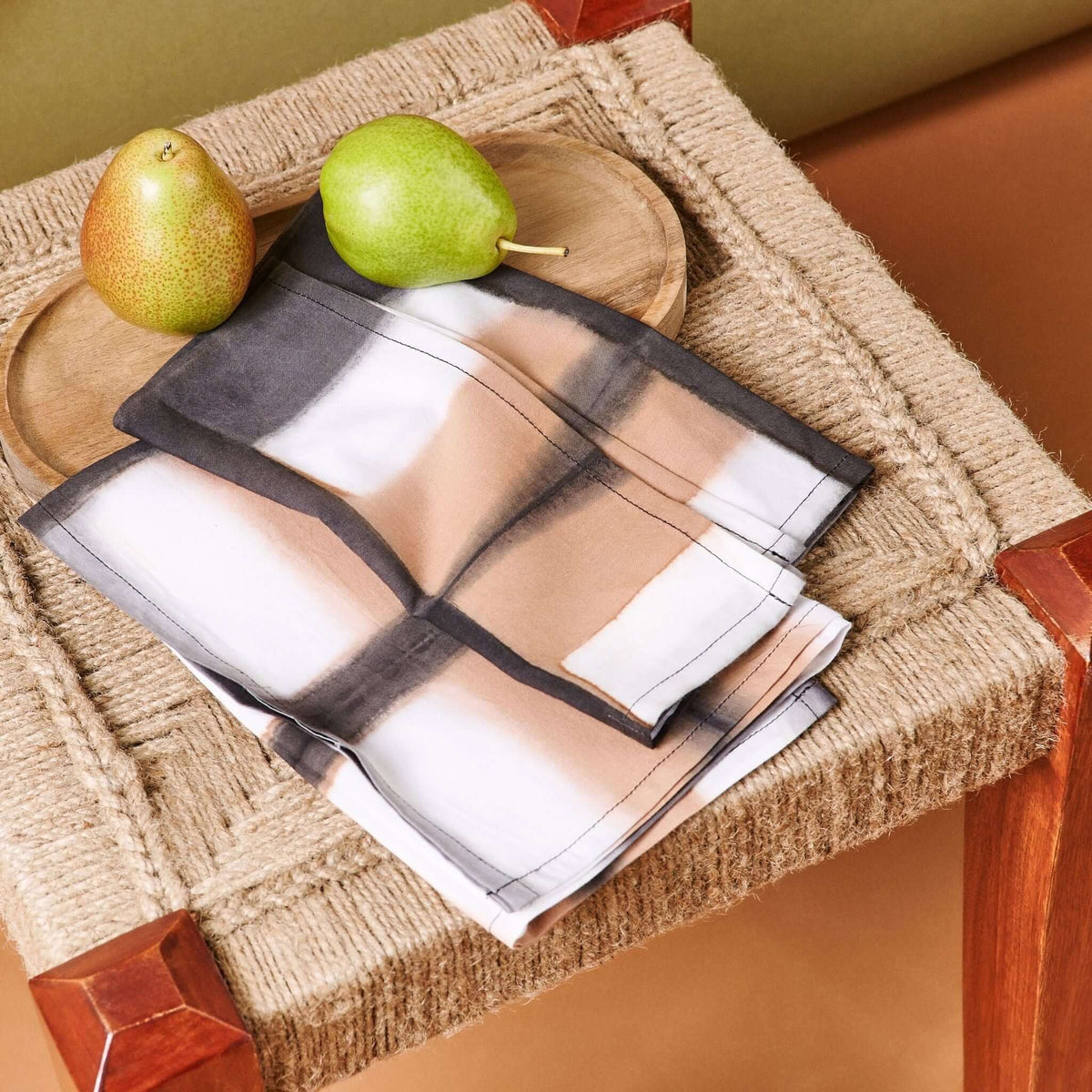 Variously Chowk Black cotton napkins folded on a woven stool beside green pears, showing the hand-dyed black, tan and white grid pattern