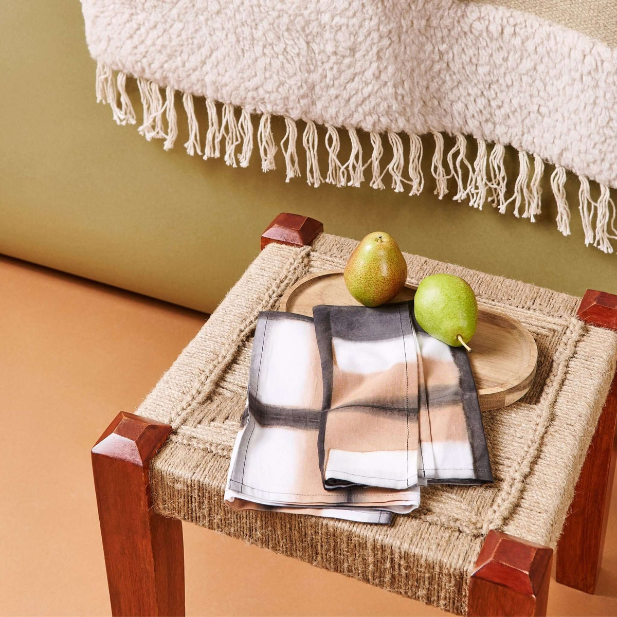 Chowk Black napkin set styled with green pears on a woven stool beneath a fringed neutral textile