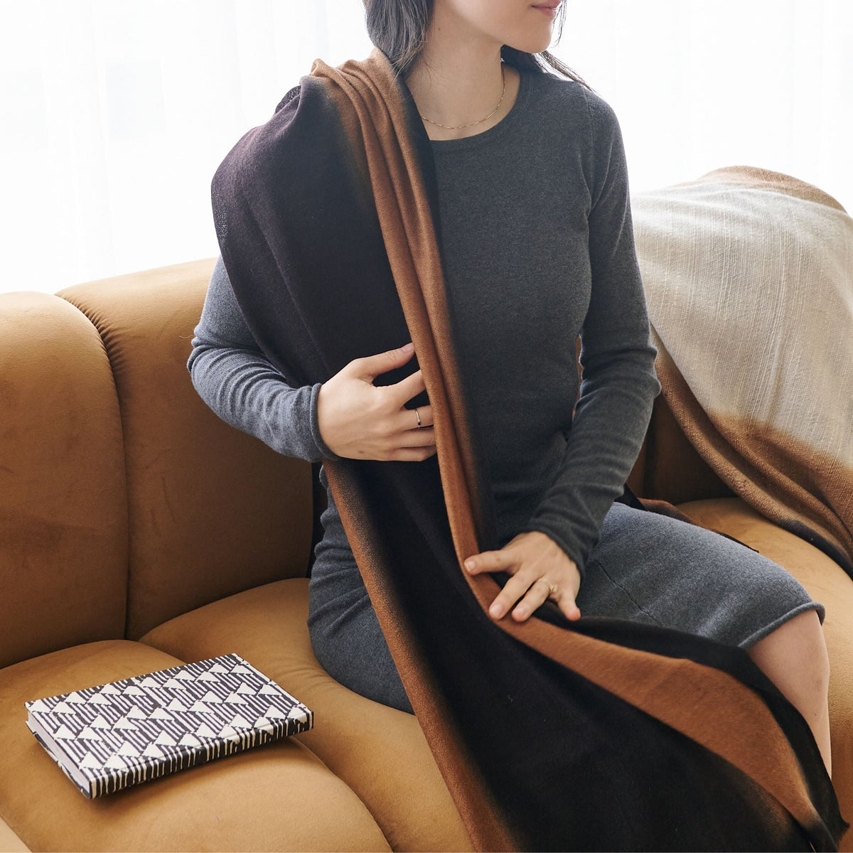 Woman seated beside the Tria journal on a tan sofa, wearing a dark dress with a wide brown shoulder strap