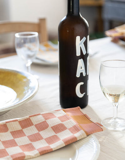 Coral-and-cream Tortuga Forma Dinner Napkins Double Sided Checker - Quiet Season folded across a white plate on a dining table, beside a dark bottle with large white lettering, wine glasses and other tableware.