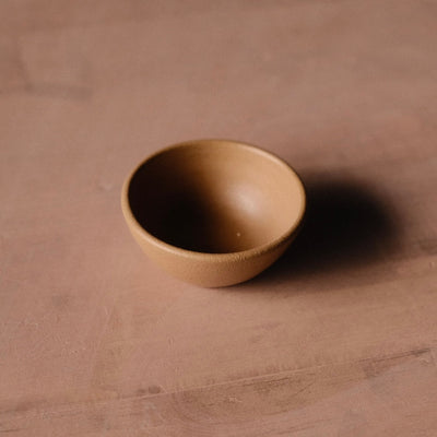 Golden-tan Settle Ceramics Wander Tiny Bowl viewed at an angle on a warm brown tabletop, showing its rounded ceramic body, gently flared rim and finely speckled glaze, with a dark shadow to the right.