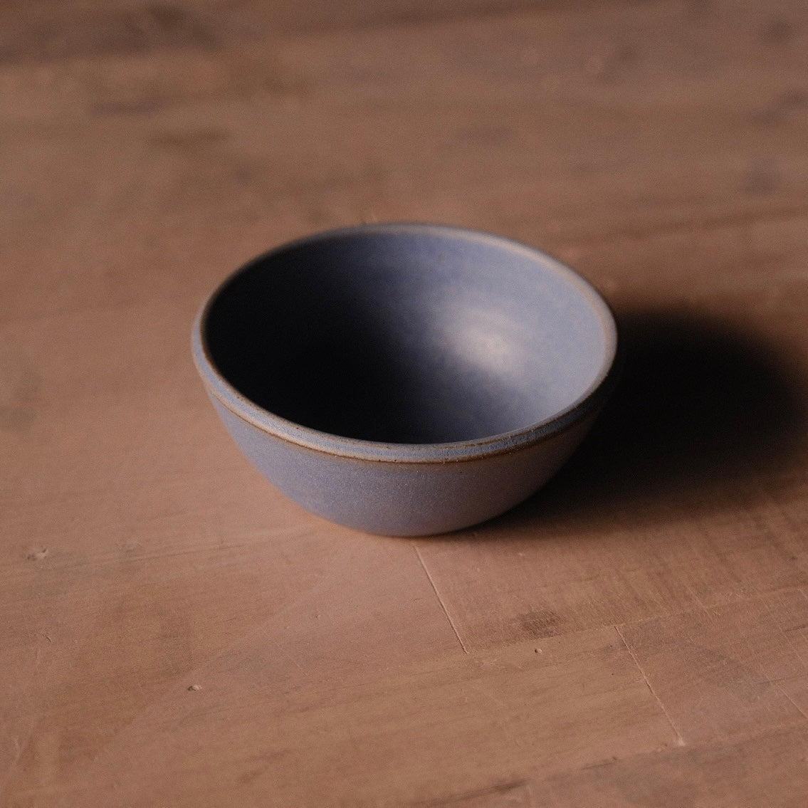 Grey Settle Ceramics Wander Side Bowl viewed at an angle on a warm brown tabletop, showing its rounded ceramic body, gently flared sides and thin contrasting brown rim, with a deep shadow to the right.