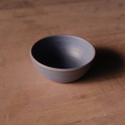 Grey Settle Ceramics Wander Side Bowl viewed at an angle on a warm brown tabletop, showing its rounded ceramic body, gently flared sides and thin contrasting brown rim, with a deep shadow to the right.
