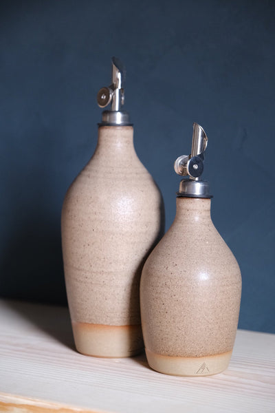 Speckled beige Settle Ceramics Wander Oil Bottle beside a taller matching bottle on pale wood against a dark blue backdrop, with fine horizontal surface lines, metal pouring spouts and cream bases.