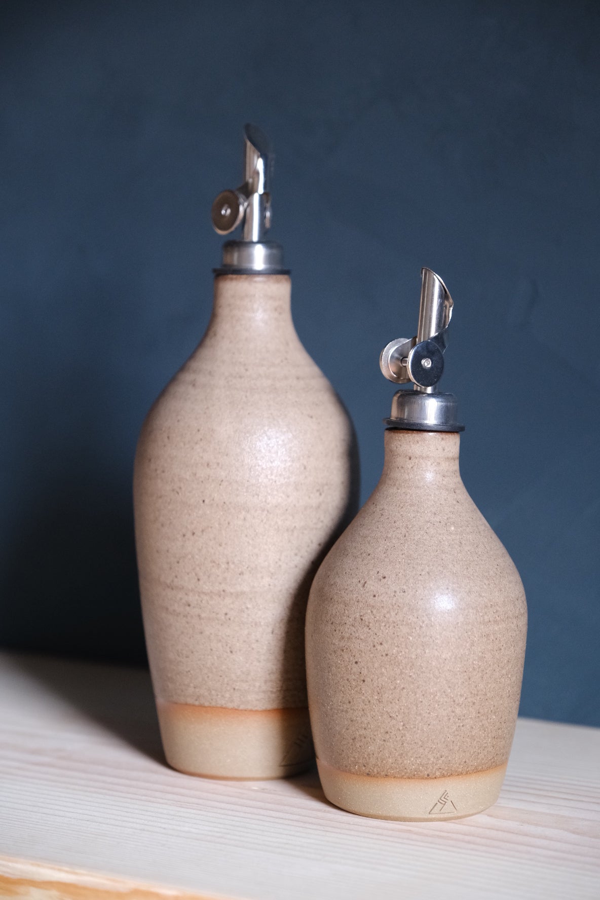 Pale speckled beige Settle Ceramics Wander Tall Oil Bottle beside a shorter matching bottle on a light wooden surface, showing gently ridged ceramic bodies and silver-toned pourers against dark blue.