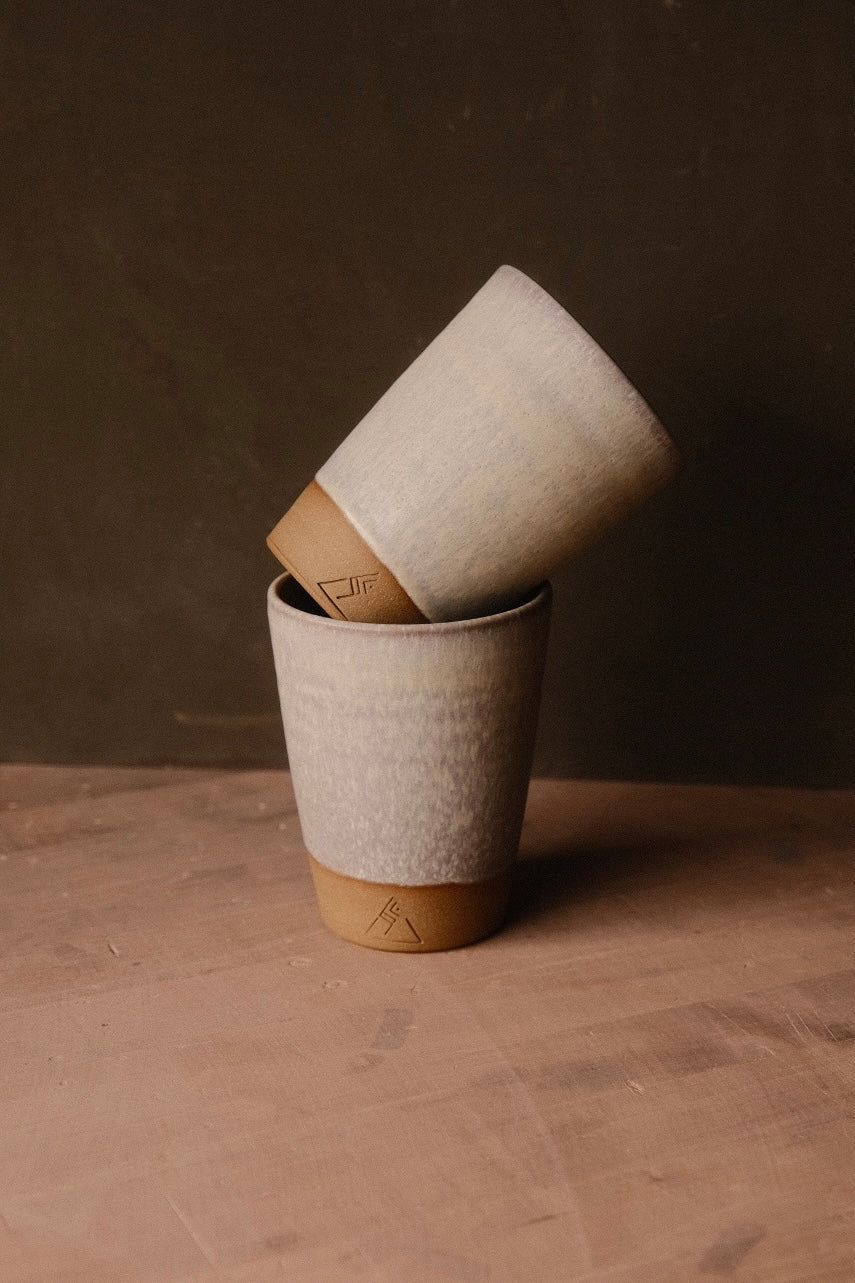Two cream Settle Ceramics Wander Tall Tumblers on a brown tabletop, one upright and the other tilted inside its rim. The handleless ceramic cups taper toward ochre bases marked with small geometric impressions, with softly mottled glaze and subtle horizontal surface lines.