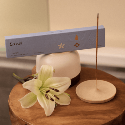 Looshi Change incense box displayed beside a white lily and a burning incense stick in a round holder on a wooden table