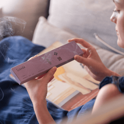 Seated person holding and examining a lavender box of Looshi Allure incense sticks above a table with books and papers
