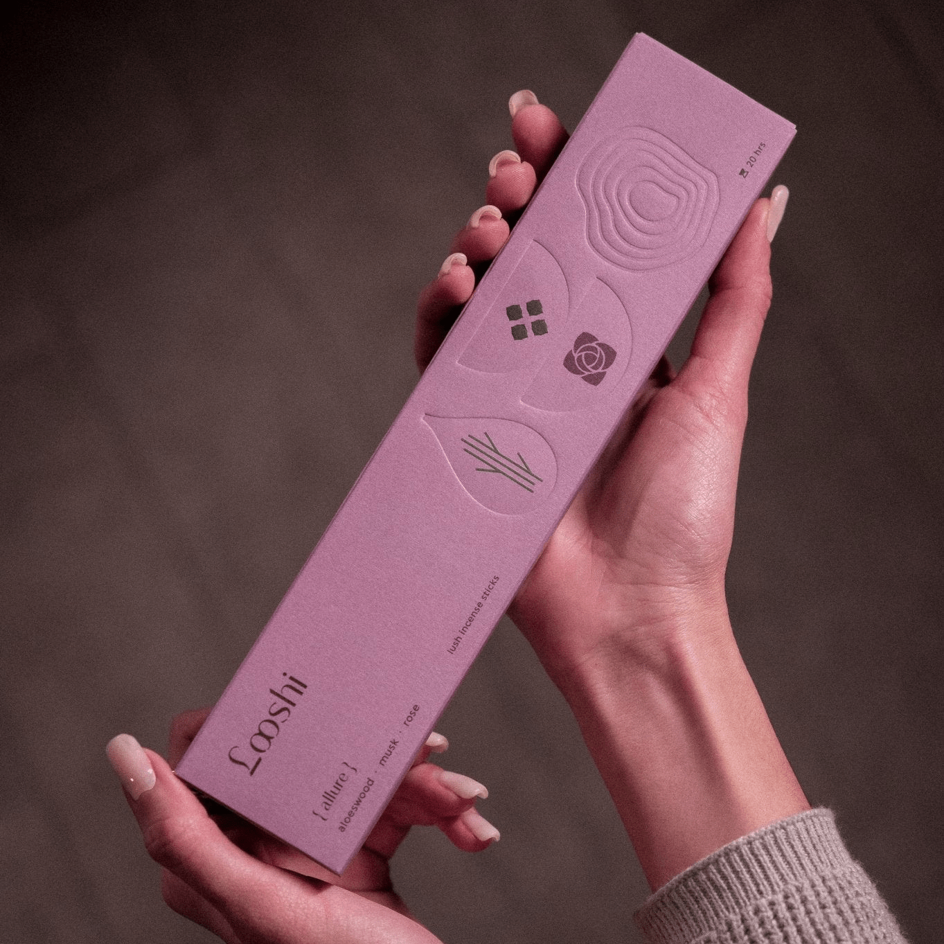 Two hands holding a tall lavender box of Looshi Allure incense sticks with embossed decorative designs against a dark brown background