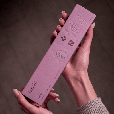 Two hands holding a tall lavender box of Looshi Allure incense sticks with embossed decorative designs against a dark brown background